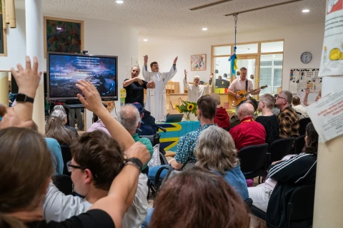 Menschen sitzen in einem Raum mit einer erh&ouml;hten B&uuml;hne. Auf der B&uuml;hne stehen mehrere Personen, darunter zwei in wei&szlig;en Gew&auml;ndern mit erhobenen Armen und ein Mann mit Akustikgitarre. Im Vordergrund heben Mitglieder des Publikums ihre H&auml;nde. Links ein Monitor mit dem eingeblendeten Text einer Liedstrophe. Im Raum befinden sich Fenster, ein Wandbild und eine Uhr an der Wand.