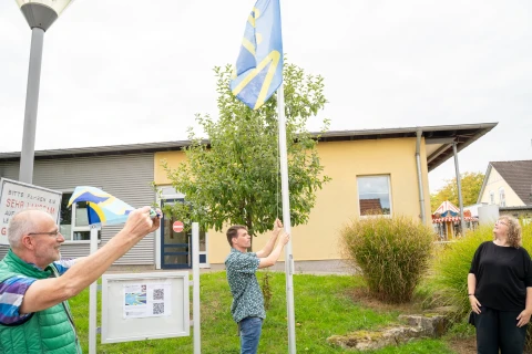 Drei Personen auf einer Gr&uuml;nfl&auml;che vor einem Geb&auml;ude mit gelber und grauer Fassade. Eine Person in der Mitte hisst eine blau-gelbe Flagge an einem Mast. Links fotografiert ein Mann mit Brille das Geschehen, rechts steht eine Frau und blickt zur Flagge. Ein kleiner Baum, ein Info-Display und ein Strauch sind im Hintergrund zu sehen.