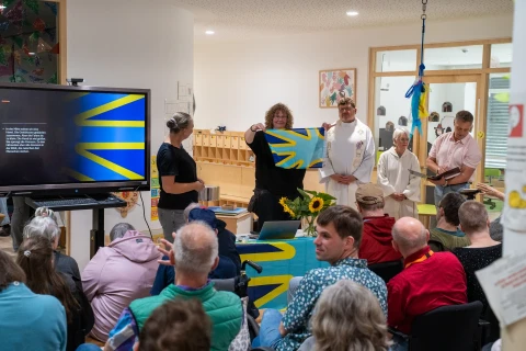 Menschen sitzen vor einer Pr&auml;sentation in einem Raum. Im Vordergrund Publikum auf St&uuml;hlen, einige stehen. Links ein Bildschirm mit einer gelb-blauen Grafik. In der Mitte halten zwei Personen eine gelb-blaue Flagge hoch, hinter einem Tisch mit Sonnenblumen. Weitere Personen rechts daneben. Holzm&ouml;bel und eine T&uuml;r im Hintergrund.