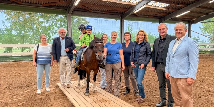 Gruppe von neun Erwachsenen in einer Reithalle, die um ein Pony mit einem Kind stehen; das Kind tr&auml;gt einen gr&uuml;nen Pullover und einen blauen Helm, sitzt auf dem Pony und wird von einem Mann gef&uuml;hrt, Pony und Kind stehen auf einem h&ouml;lzernen Steg, Hintergrund gr&uuml;ne B&auml;ume und Lichtdurchl&auml;ssige W&auml;nde.