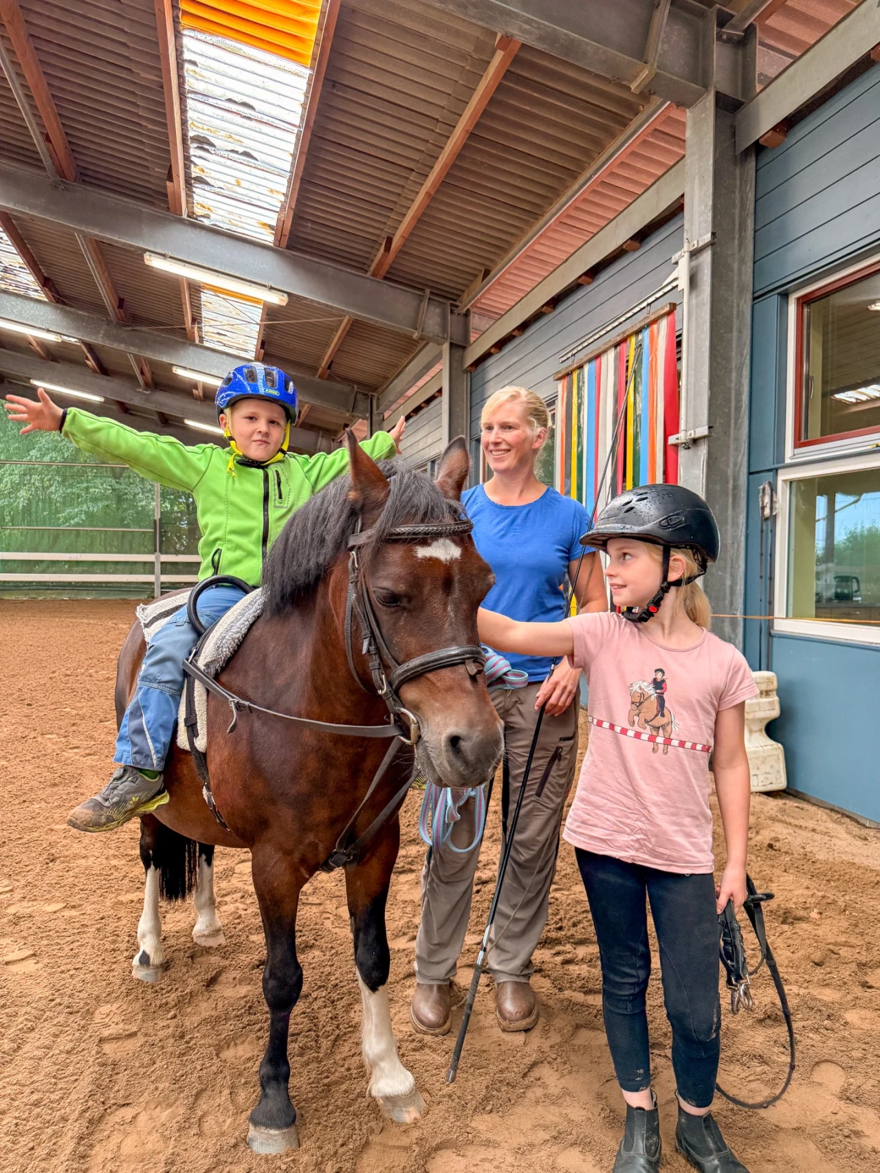 Ein kleiner Junge mit gr&uuml;nem Hoodie und blauer Reitkappe sitzt auf einem braunen Pony mit wei&szlig;er Blesse und Zaumzeug, Arme ausgestreckt. Neben dem Pony stehen eine Frau in blauem Shirt und eine junge Reiterin in rosa T-Shirt mit schwarzer Reitkappe, die das Pony streichelt. Innenraum einer Reithalle mit sandigem Boden und bunten Streifen an einer Wand.
