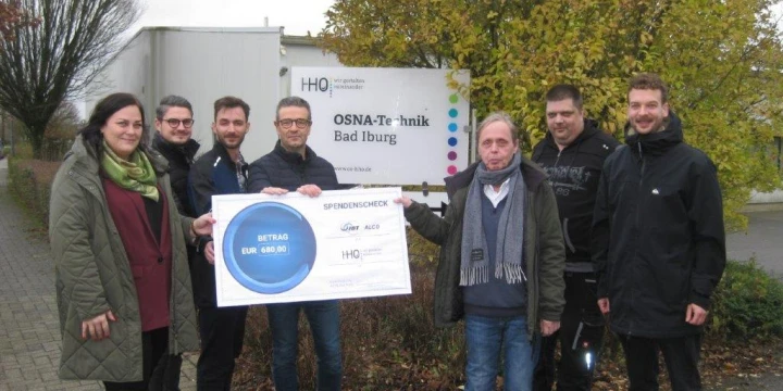 Mehrere Personen stehen vor einem Schild mit der Aufschrift "OSNA-Technik Bad Iburg" und halten gemeinsam einen gro&szlig;en symbolischen Spendenscheck mit einem Betrag von 680,80 Euro. Im Hintergrund herbstlicher Baum und Containergeb&auml;ude.