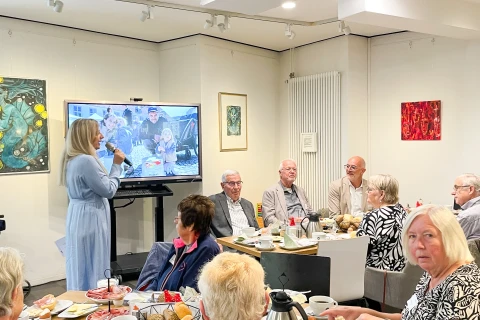 Frau in hellblauem Kleid spricht mit Mikrofon vor einer Gruppe &auml;lterer Menschen, die an einem Tisch mit Geschirr, Kaffee und Fr&uuml;hst&uuml;cksgeb&auml;ck sitzen. Im Hintergrund ein gro&szlig;er Bildschirm mit einem Foto, W&auml;nde mit farbigen Gem&auml;lden und ein Heizk&ouml;rper.