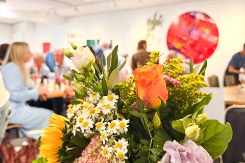 Blumenstrau&szlig; in Glasvase mit Sonnenblumen, wei&szlig;en G&auml;nsebl&uuml;mchen, orangefarbener Rose und lila Bl&uuml;te im Vordergrund; unscharf im Hintergrund mehrere sitzende Personen und helle Wand mit abstrakten Kunstwerken, darunter ein gro&szlig;es rotes Rundbild.