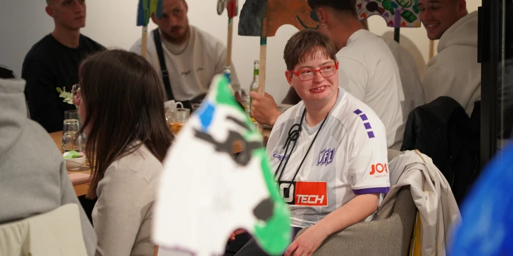 Personen sitzen an einem Tisch in einem geschlossenen Raum, auf dem Tisch stehen Gl&auml;ser und Dekorationen. Im Vordergrund ist ein dekoratives Papiermotiv in Form eines Pferdekopfes an einem Stab sichtbar. Im Hintergrund halten mehrere Personen bunte, bemalte Papierfiguren in &auml;hnlicher Form. Eine Person tr&auml;gt ein wei&szlig;-lilanes Trikot.