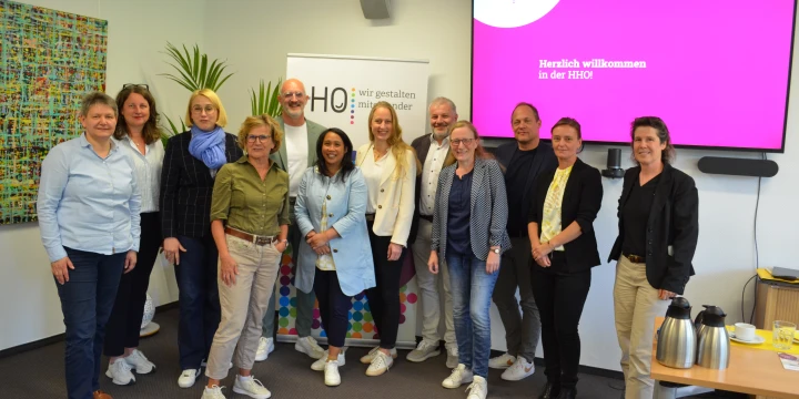 Gruppe von zehn Personen in einem Konferenzraum, stehend vor einer Pr&auml;sentationsfl&auml;che. Rechts ein Bildschirm mit pinkem Hintergrund und der Aufschrift "Herzlich willkommen in der HHO". Links ein Roll-Up mit der Aufschrift "HHO &ndash; wir gestalten inklusive Vielfalt". Tisch rechts vorne mit Getr&auml;nkekaraffen, Gl&auml;sern und Tassen.