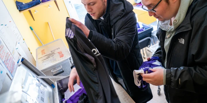 Zwei Personen sortieren Textilien. Links h&auml;lt eine Person ein schwarzes und lilafarbenes Kleidungsst&uuml;ck mit Schriftzug hoch. Rechts h&auml;lt eine Person &auml;hnliche Kleidung. Im Hintergrund blaue Regale, Reinigungsger&auml;te und Kisten.