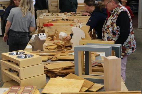 Personen betrachten an einem langen Tisch verschiedene Holzarbeiten, darunter dekorative Objekte, Stapel von Holzplatten und Holzfiguren. Im Hintergrund wei&szlig;e W&auml;nde mit einem Regal, einer Uhr und einer gelben Aufh&auml;ngung von der Decke.