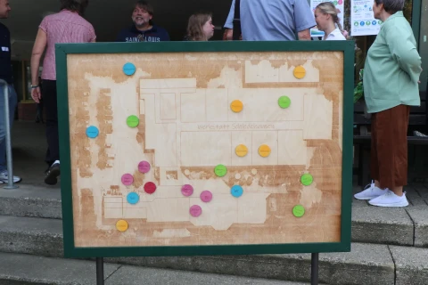 Holztafel mit eingraviertem Grundriss und der Aufschrift "Werkstatt Schiefbahnen". Auf dem Grundriss sind bunte, runde Magnete in Blau, Pink, Gelb und Gr&uuml;n verteilt. Im Hintergrund stehen mehrere Personen in Freizeitkleidung vor einem Geb&auml;ude.