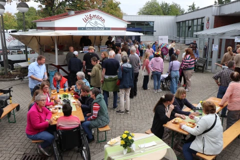 Menschen sitzen an Holzb&auml;nken und Tischen und essen oder unterhalten sich. Im Hintergrund befindet sich ein wei&szlig;er Imbisswagen mit der Aufschrift &bdquo;Wi&szlig;mann Catering und Eventservice&ldquo;. Rundherum stehen weitere Personen in Gruppen zusammen, einige in der Essensausgabe-Schlange. Gelb bl&uuml;hende Blumen stehen auf den Tischen. Geb&auml;ude mit Glas- und Metallfassaden umgeben den Platz.