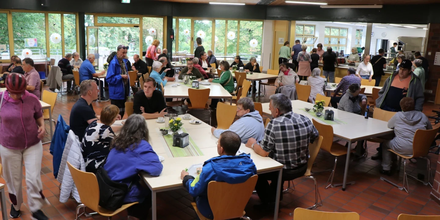 Menschen sitzen und stehen in einer Kantine mit gro&szlig;en Fenstern, durch die gr&uuml;ne B&auml;ume sichtbar sind. Gelbe Holzst&uuml;hle stehen an Tischen, die mit gr&uuml;nen Tischl&auml;ufern und kleinen Pflanzen dekoriert sind. Im Hintergrund Selbstbedienungsbereich mit Essensausgabe, moderner Deckenbeleuchtung und Terrakottaboden.
