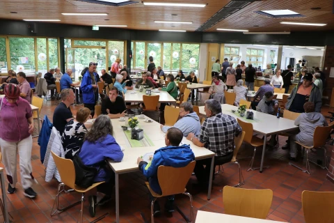 Menschen sitzen und stehen in einer Kantine mit gro&szlig;en Fenstern, durch die gr&uuml;ne B&auml;ume sichtbar sind. Gelbe Holzst&uuml;hle stehen an Tischen, die mit gr&uuml;nen Tischl&auml;ufern und kleinen Pflanzen dekoriert sind. Im Hintergrund Selbstbedienungsbereich mit Essensausgabe, moderner Deckenbeleuchtung und Terrakottaboden.