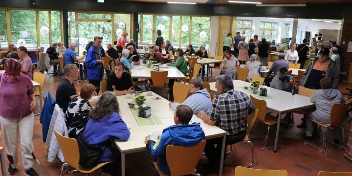 Menschen sitzen und stehen in einer Kantine mit gro&szlig;en Fenstern, durch die gr&uuml;ne B&auml;ume sichtbar sind. Gelbe Holzst&uuml;hle stehen an Tischen, die mit gr&uuml;nen Tischl&auml;ufern und kleinen Pflanzen dekoriert sind. Im Hintergrund Selbstbedienungsbereich mit Essensausgabe, moderner Deckenbeleuchtung und Terrakottaboden.