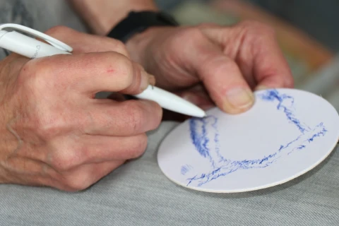 H&auml;nde einer Person, die mit einem wei&szlig;en Kugelschreiber auf eine runde, wei&szlig;e Pappscheibe skizziert; das Motiv zeigt grob gezeichnete blaue Linien, die eine Landschaft oder abstrakte Form andeuten. Im Hintergrund ein unscharfer Stoffuntergrund und ein dunkles Uhrenarmband am Handgelenk.