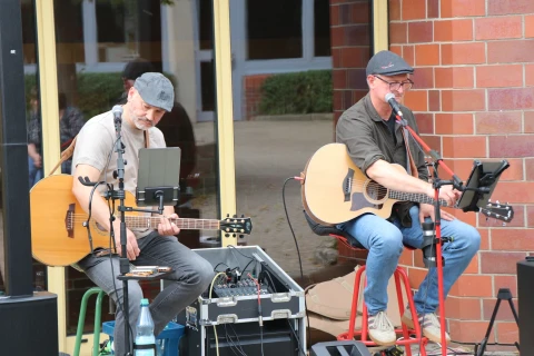 Zwei M&auml;nner mit Schirmm&uuml;tzen spielen Akustikgitarren und singen in Mikrofone. Links liegt ein Mischpult auf einem Koffer, rechts ein Tablet auf einem St&auml;nder. Sie sitzen vor einer Glasfront mit gelbem Rahmen und einer roten Backsteinwand. Davor steht eine Wasserflasche.