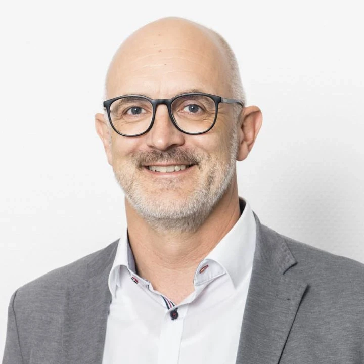 Glatzk&ouml;pfiger Mann mittleren Alters mit Brille und grauem Bart, tr&auml;gt ein graues Sakko &uuml;ber einem wei&szlig;en Hemd mit gemustertem Kragen, l&auml;chelt freundlich vor einem wei&szlig;en Hintergrund.