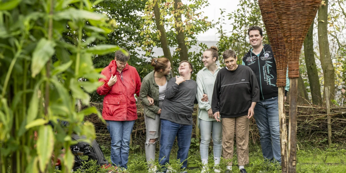 Sechs Personen unterschiedlichen Alters stehen auf einer gr&uuml;nen Wiese; sie lachen und interagieren miteinander. Eine Person tr&auml;gt eine rote Jacke, andere tragen legere Kleidung. Im Hintergrund sind B&auml;ume, Str&auml;ucher und ein geflochtenes Weidenelement am rechten Bildrand sichtbar.