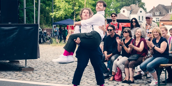 Eine Person in einem wei&szlig;en Hemd hebt eine andere Person in dunkler Hose und wei&szlig;en Schuhen, die pinke Socken tr&auml;gt. Im Hintergrund sitzen mehrere Menschen auf Holzb&auml;nken, einige applaudieren. Pflastersteine, eine B&uuml;hne und eine Gr&uuml;nfl&auml;che mit B&auml;umen sind sichtbar, daneben Zelte und Geb&auml;ude.