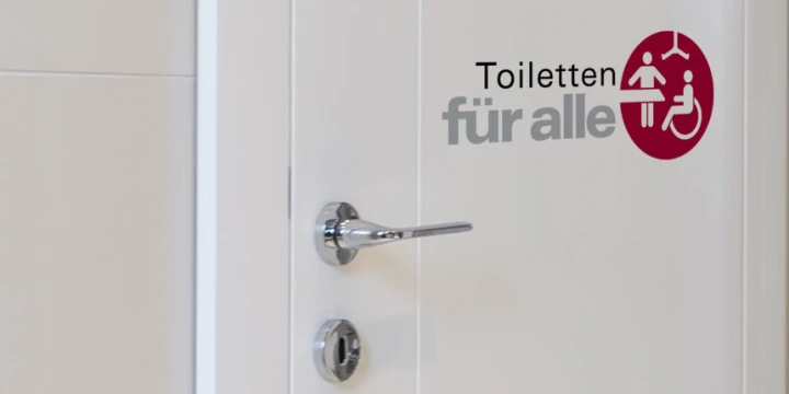 Wei&szlig;e T&uuml;r mit silbernem T&uuml;rgriff und Schlie&szlig;mechanismus. Auf der T&uuml;r Schriftzug "Toiletten f&uuml;r alle" in schwarzer und grauer Schrift, daneben ein roter Kreis mit wei&szlig;en Symbolen f&uuml;r Personen, Kinder und Rollstuhlfahrer.