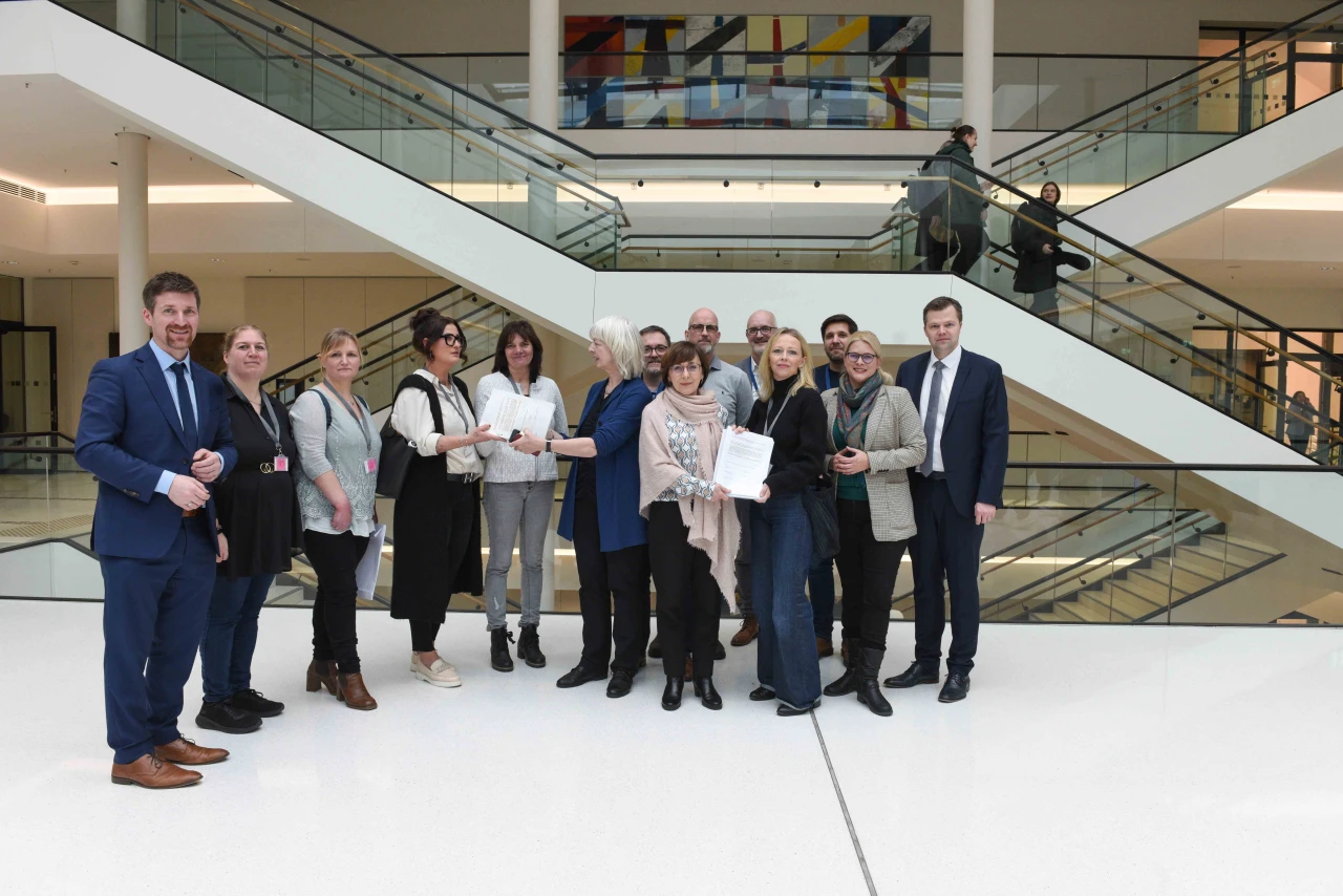 Gruppe von elf Personen in einem hellen, modernen Geb&auml;ude mit wei&szlig;en W&auml;nden und Glasgel&auml;ndern, in der Mitte eine &Uuml;bergabe von Papieren. Im Hintergrund zwei Treppenaufg&auml;nge und eine bunte Wanddekoration.