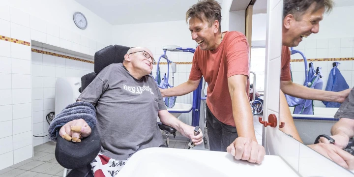Ein Mann mit einer Beeintr&auml;chtigung der im Rollstuhl ist und ein weiterer Mann der sich mit ihm austauscht. Beide l&auml;cheln.