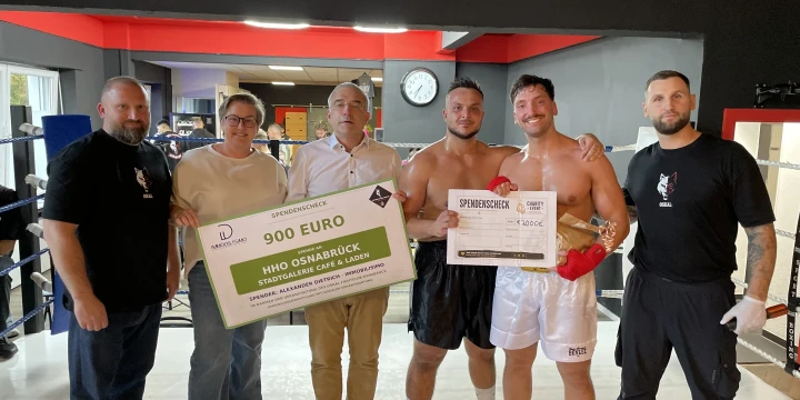 Sechs Personen stehen in einem Raum mit Boxringen. Zwei Personen in der Mitte tragen Boxshorts und Boxhandschuhe. Sie halten zwei gro&szlig;e Spendenchecks: Links ein gr&uuml;ner Check mit "900 EURO" an IHO Osnabr&uuml;ck, rechts ein wei&szlig;er Check mit "2.300&euro;". Im Hintergrund schwarze und rote W&auml;nde, eine Uhr und Trainingsger&auml;te.
