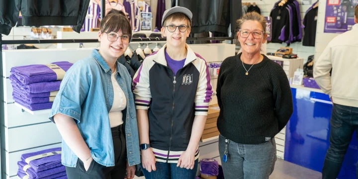 Drei Personen stehen nebeneinander in einem Gesch&auml;ft mit Verkaufsartikeln. Links Person mit blauer Jeansjacke und heller Hose, in der Mitte Person mit lila-baseballartigem Oberteil und Kappe, rechts Person in schwarzem Pullover und grauer Hose. Regale mit lilafarbenen Handt&uuml;chern und Kleidungsst&uuml;cken im Hintergrund, Preisschilder und Werbeschilder an den W&auml;nden.