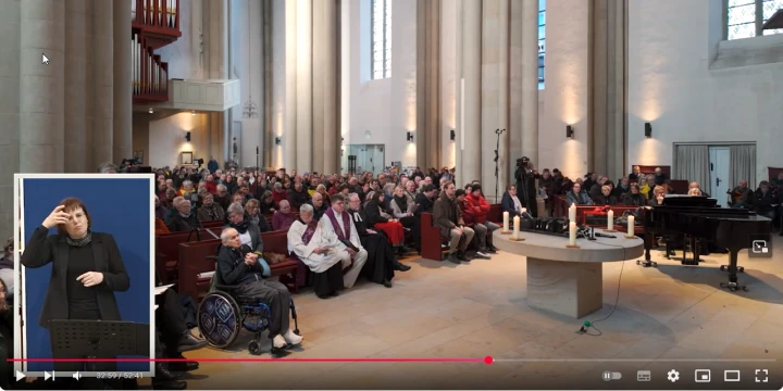 Eine gro&szlig;e Kirche mit hohen S&auml;ulen und Buntglasfenstern, gef&uuml;llt mit vielen sitzenden Menschen auf Holzb&auml;nken. Im Vordergrund ein runder Altartisch mit Kerzen und Dekorationen, rechts daneben ein schwarzer Fl&uuml;gel. Links im Bild ist ein Bereich f&uuml;r Geb&auml;rdendolmetscher eingeblendet.