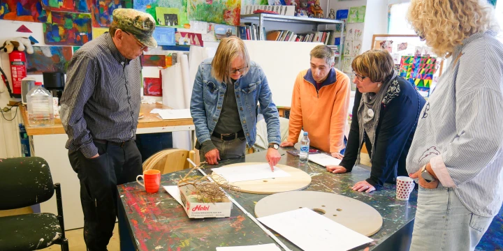 Sechs Personen stehen um einen schwarzen, bunt beklecksten Holztisch in einem Kunst- oder Kreativraum. Auf dem Tisch liegen runde Holzscheiben, Papierbl&auml;tter, eine Flasche Wasser, Tassen und eine Verpackung. Im Hintergrund bunt bemalte Bilder an W&auml;nden und Regale mit k&uuml;nstlerischen Utensilien.