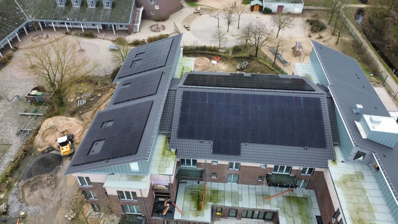 Luftaufnahme eines Geb&auml;udekomplexes mit dunklen Solarpaneelen auf den Dachfl&auml;chen. Das Geb&auml;ude besteht aus mehreren miteinander verbundenen Fl&uuml;geln aus rotem Ziegelstein mit grauen D&auml;chern und gr&uuml;nen Glasdetails. Links unten Baustellenbereich mit gelbem Bagger. Umgebung umfasst B&auml;ume, Wege und angrenzende Geb&auml;ude.