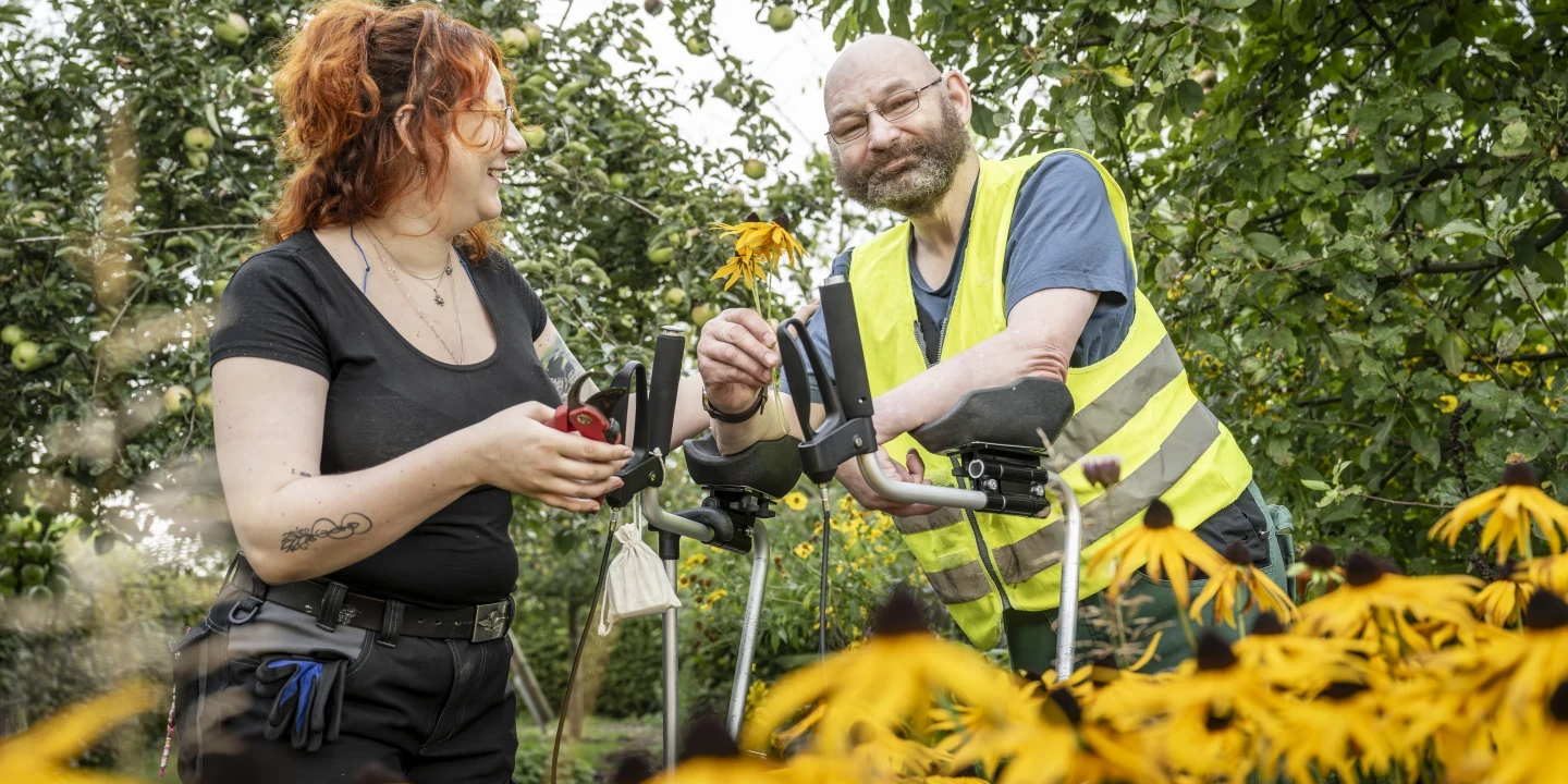 Frau mit roten Haaren, schwarzem T-Shirt und Schere in der Hand steht neben einem Mann mit Glatze, Vollbart, gelber Warnweste und Unterarmgehst&uuml;tzen. Beide betrachten eine gelbe Blume, umgeben von weiteren gelben Blumen und gr&uuml;nem Laub im Freien.