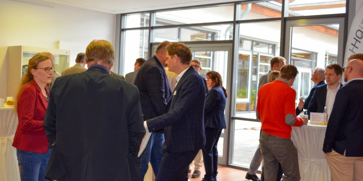 Personengruppe in einem hellen Raum mit gro&szlig;en Fenstern und Blick auf einen Innenhof. Stehende Menschen in Anz&uuml;gen und Casual-Bekleidung unterhalten sich an mehreren Stehtischen mit wei&szlig;en Hussen. Ein transparenter Aufsteller mit Schriftzug "HQH" steht rechts im Bild.