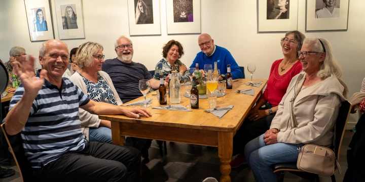 Mehrere Personen sitzen an einem rechteckigen Holztisch in einem Raum mit heller Beleuchtung. Auf dem Tisch stehen Flaschen, Gl&auml;ser und Servietten. Im Hintergrund befinden sich an einer wei&szlig; gestrichenen Wand gerahmte Schwarz-Wei&szlig;-Fotografien in einer Reihe.