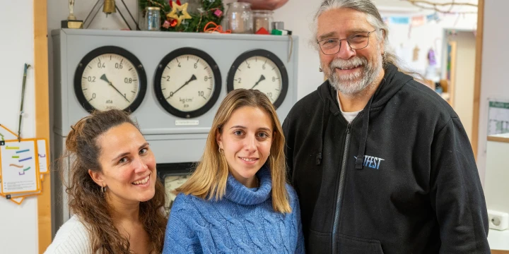 Eine Gruppe von drei Personen in einem Raum mit wei&szlig;er Wand und verschiedenen Gegenst&auml;nden. Im Hintergrund eine graue Schaltanlage mit vier analogen Anzeigen und einem darauf platzierten weihnachtlich dekorierten Kranz, einer rosafarbenen Kugel und Glasbeh&auml;ltern. Links h&auml;ngt ein Papier mit einer bunten Grafik an der Wand.