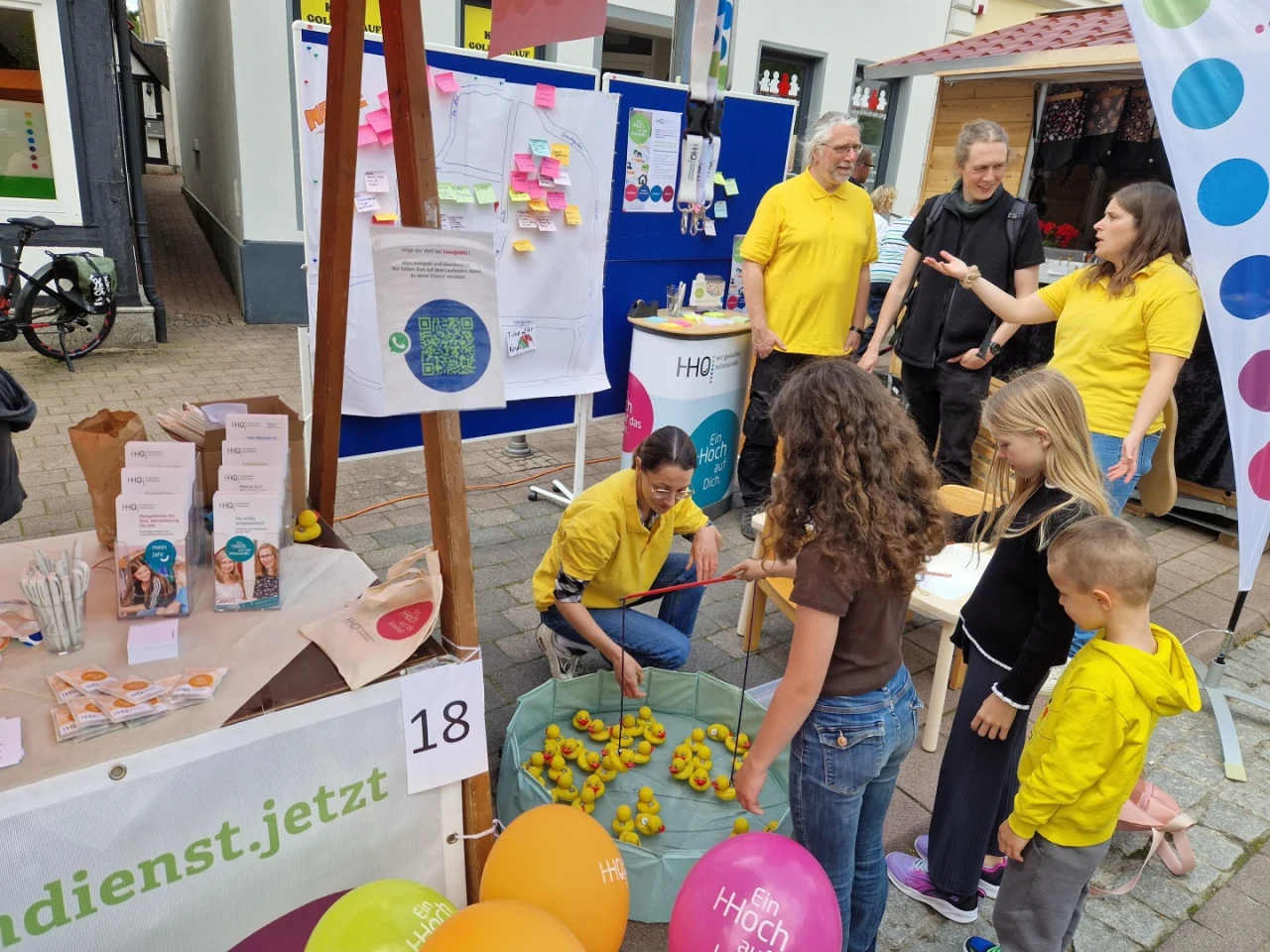 Mehrere Personen an einem bunten Informationsstand auf einer Veranstaltung. Links ein Holztisch mit Brosch&uuml;ren, T&uuml;ten und Spielzeugenten auf einem gr&uuml;nen Tuch. Ein Mann kniet und ordnet Enten, w&auml;hrend Kinder zuschauen. Im Hintergrund Pinnw&auml;nde mit Notizen und zwei Personen in gelben Shirts, rechts eine Frau in Interaktion mit Besuchern. Ballons mit Aufschriften (&bdquo;Bin hoch&ldquo;) h&auml;ngen an der Standecke, Beschriftung &bdquo;18&ldquo; sichtbar. Stadtumgebung im Hintergrund.