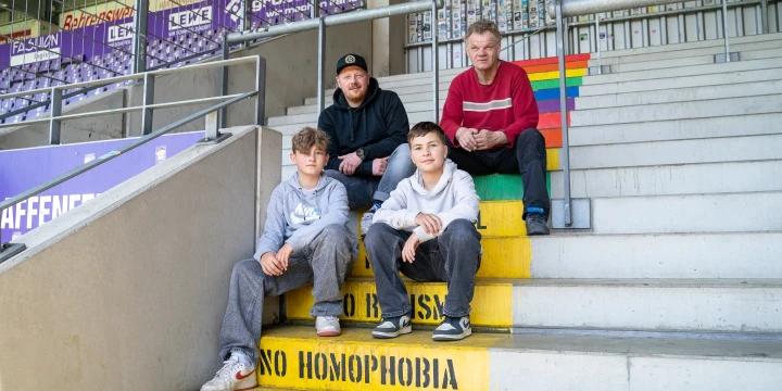Vier sitzende Personen auf einer Betontreppe in einem Stadion. Die Stufen tragen gelbe Markierungen mit den Schriftz&uuml;gen "NO HOMOPHOBIA" und "NO VIOLENCE". Rechts auf der Treppe ist eine Regenbogenflagge gemalt. Im Hintergrund sind lila Werbebanner mit Text und eine Wand mit Stickern sichtbar.
