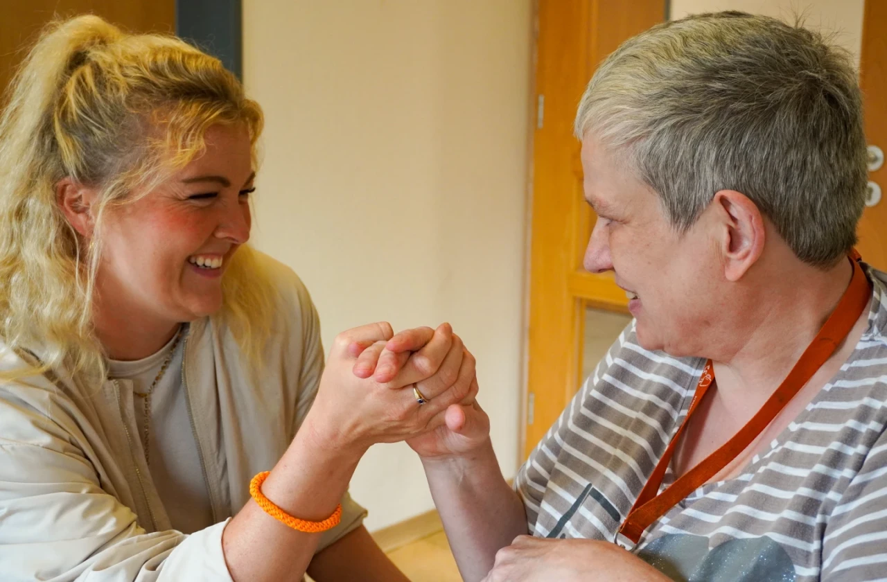 Zwei Personen in einem Innenraum, die einander die H&auml;nde reichen. Links eine Person mit blondem Haar, beigem Oberteil und orangefarbenem Armband, die nach rechts blickt. Rechts eine Person mit grauem kurzhaarigem Haar, gestreiftem Oberteil und Brillenkette, die nach links blickt. Im Hintergrund beiger Wandbereich und eine T&uuml;r.