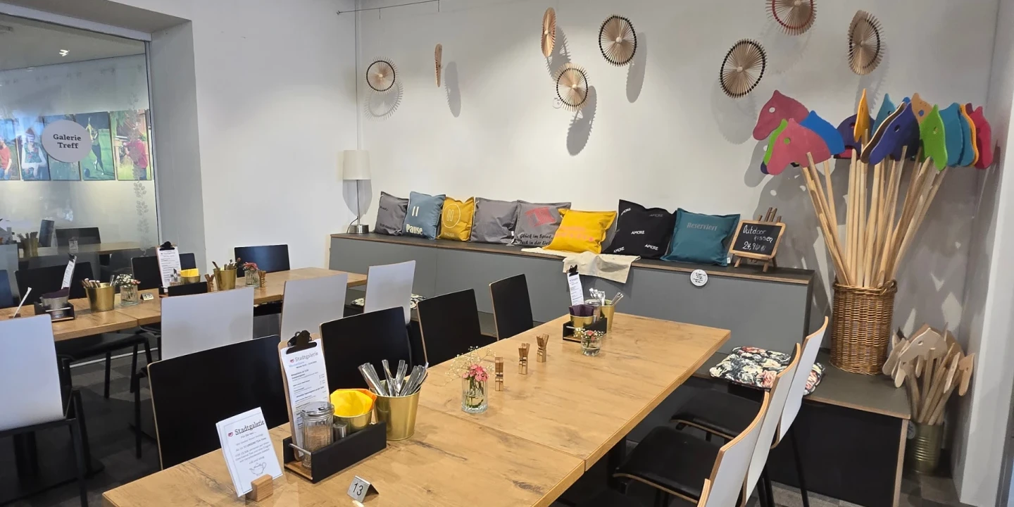 Holztische mit Dekorationen wie kleine Glasvasen und Men&uuml;karten in einem Caf&eacute;raum. Links wei&szlig;e und schwarze St&uuml;hle, hinten eine Sitzbank mit bunten Kissen. Wanddekorationen: K&ouml;rbe und bunte Steckenpferde in einem Korb rechts.