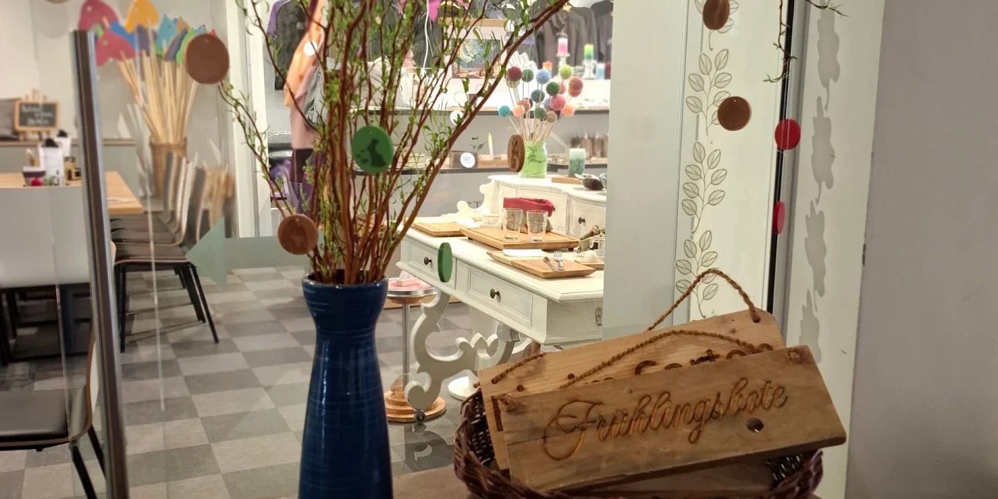 Zweig mit bunten Papierdekorationen in blauer Vase auf einem Holztisch, daneben Holzt&auml;felchen mit graviertem Schriftzug &bdquo;Fr&uuml;hlingshafe&ldquo; in ovalem Korb. Im Hintergrund ein Raum mit dekorativen Accessoires auf M&ouml;beln und Tischen, helle Bodenfliesen und Glaswand.