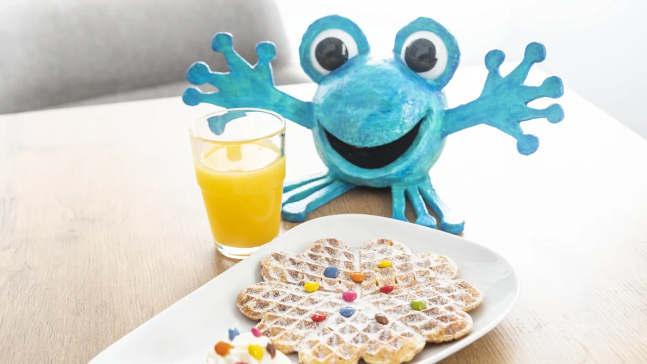 Waffel in Herzform mit bunten Zuckerdrops garniert auf wei&szlig;em ovalem Teller, daneben ein kleiner Klecks Sahne mit weiteren Zuckerdrops. Im Hintergrund ein Glas mit Orangensaft und eine blaue bastelartige Figur mit gro&szlig;en Augen, breitem L&auml;cheln und ausgestreckten Armen. Alles auf einem hellen Holztisch.