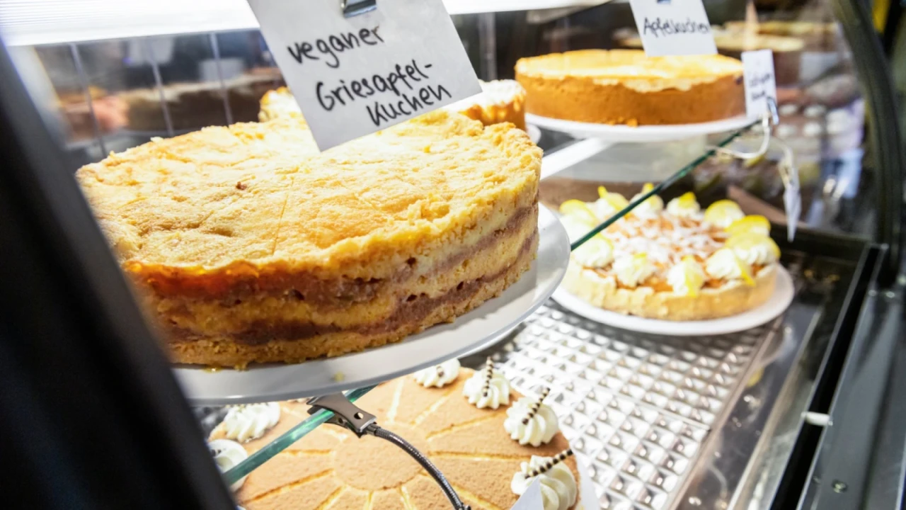 Kuchentheke mit mehreren Torten: Im Vordergrund ein veganer Grie&szlig;apfelkuchen mit goldener Kruste, rechts unten ein Kuchen mit Sahneornamenten, im Hintergrund weitere Kuchen, beschriftet mit handgeschriebenen Schildern.