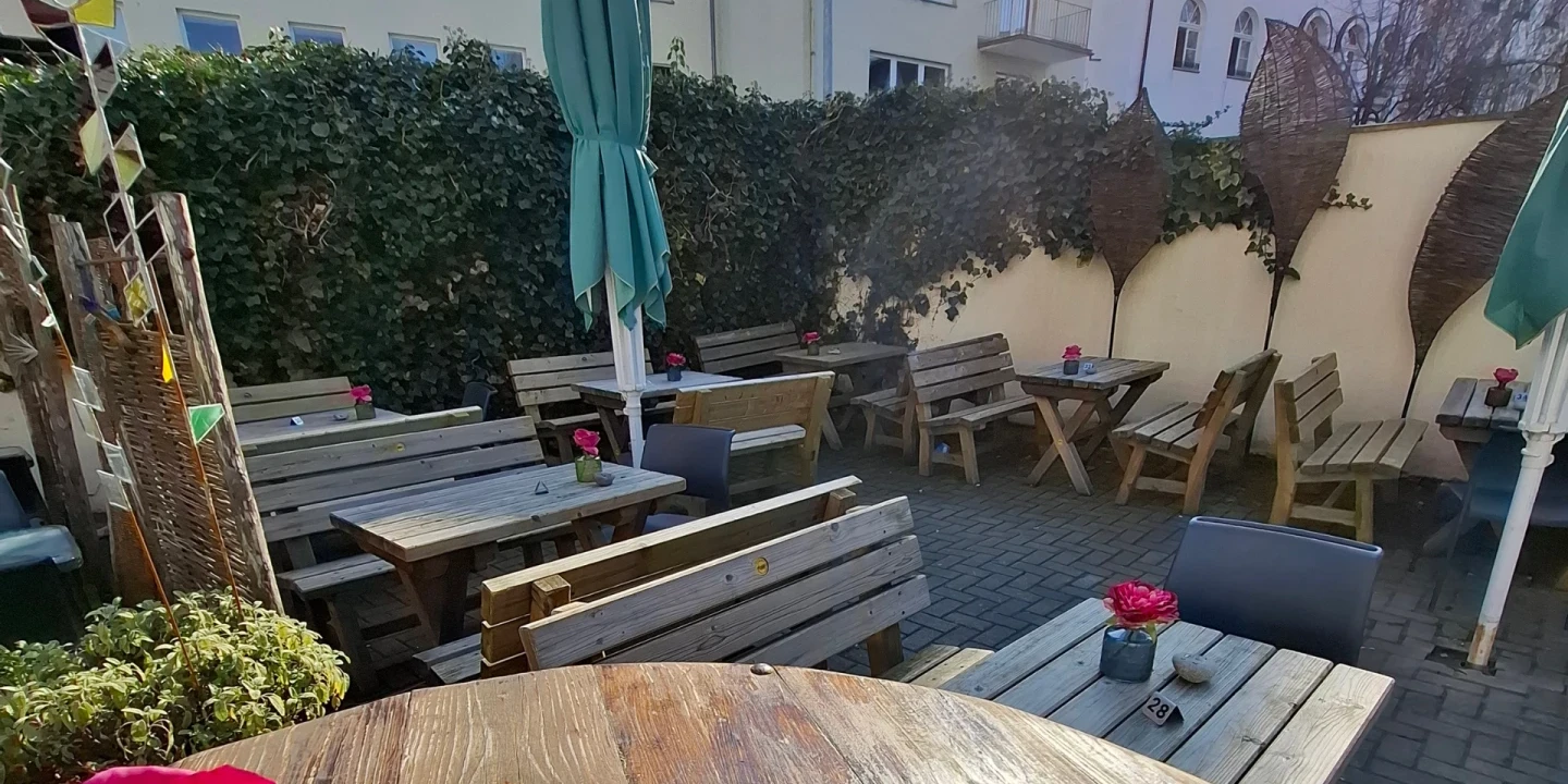 Holztische und -b&auml;nke in einem gepflasterten Innenhof, umgeben von hohen, begr&uuml;nten W&auml;nden und beige-gelben Sichtschutzw&auml;nden. Mehrere Tische haben Dekorationen wie rosa Blumen in kleinen Vasen. Zwei gro&szlig;e gr&uuml;ne Sonnenschirme sind geschlossen. Im Hintergrund sind Wohngeb&auml;ude zu sehen.
