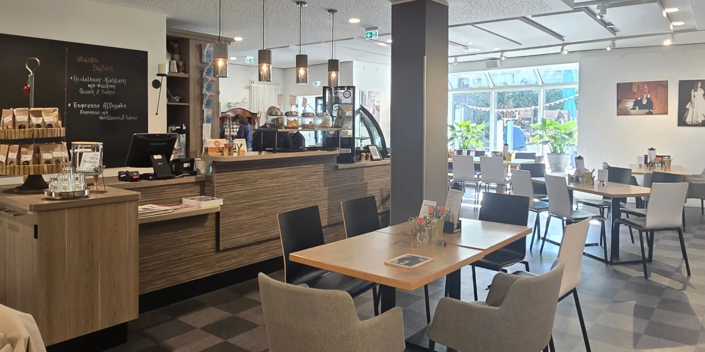Ein modern eingerichtetes Caf&eacute; mit Holzm&ouml;beln, quadratischen Tischen und gepolsterten St&uuml;hlen. Links eine Theke mit Kaffeemaschine, Display mit Geb&auml;ck und einer Tafel mit handgeschriebener Speisekarte. Helle Deckenlampen h&auml;ngen von der Decke. Rechts gro&szlig;e Fenster mit Blick auf Gr&uuml;npflanzen und Landschaft. Wand mit Bildern dekoriert.