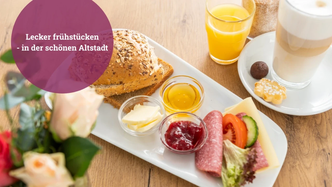 Fr&uuml;hst&uuml;ckstisch mit l&auml;nglichem wei&szlig;en Teller, darauf Mehrkornbr&ouml;tchen, kleine Gl&auml;ser mit Honig, Fruchtaufstrich und Butter, sowie Salami, K&auml;se, Gurkenscheiben, Tomate und Salat. Daneben Glas Orangensaft, Latte Macchiato, Praline, Pl&auml;tzchen. Im Vordergrund unscharfe Rosenbl&uuml;ten. Schrift auf lila Kreis: "Lecker fr&uuml;hst&uuml;cken &ndash; in der sch&ouml;nen Altstadt".