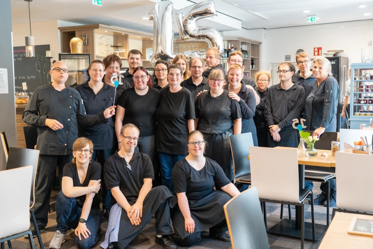 Gruppe von 15 Personen in dunkler Arbeitskleidung, stehend und kniend in einem hellen Restaurant mit gedeckten Tischen. Im Hintergrund silberne Luftballons mit der Zahl "15", Holzregale mit Geschirr und Dekoration.