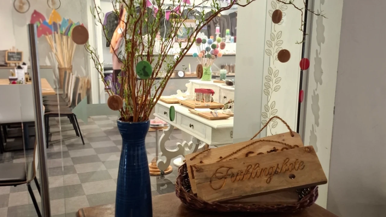 Blauer Keramikvase mit blattlosen Zweigen, an denen bunte, runde Dekorationen befestigt sind. Daneben im Korb ein Holzschild mit der Aufschrift "Fr&uuml;hlingshofe". Im Hintergrund Dekotisch mit Stoffen, bunten Ornamenten und Werkzeugen in einem hell erleuchteten Raum.
