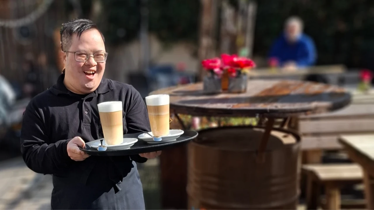 Person in schwarzer Kleidung tr&auml;gt ein Tablett mit zwei hohen Latte-Macchiato-Gl&auml;sern auf Untertassen, jeweils mit einem Kaffeel&ouml;ffel. Im Hintergrund ein runder Holztisch mit roten Blumen in einem silbernen Topf sowie unscharf andere Tische und eine sitzende Person. Outdoor-Umgebung mit Tageslicht.