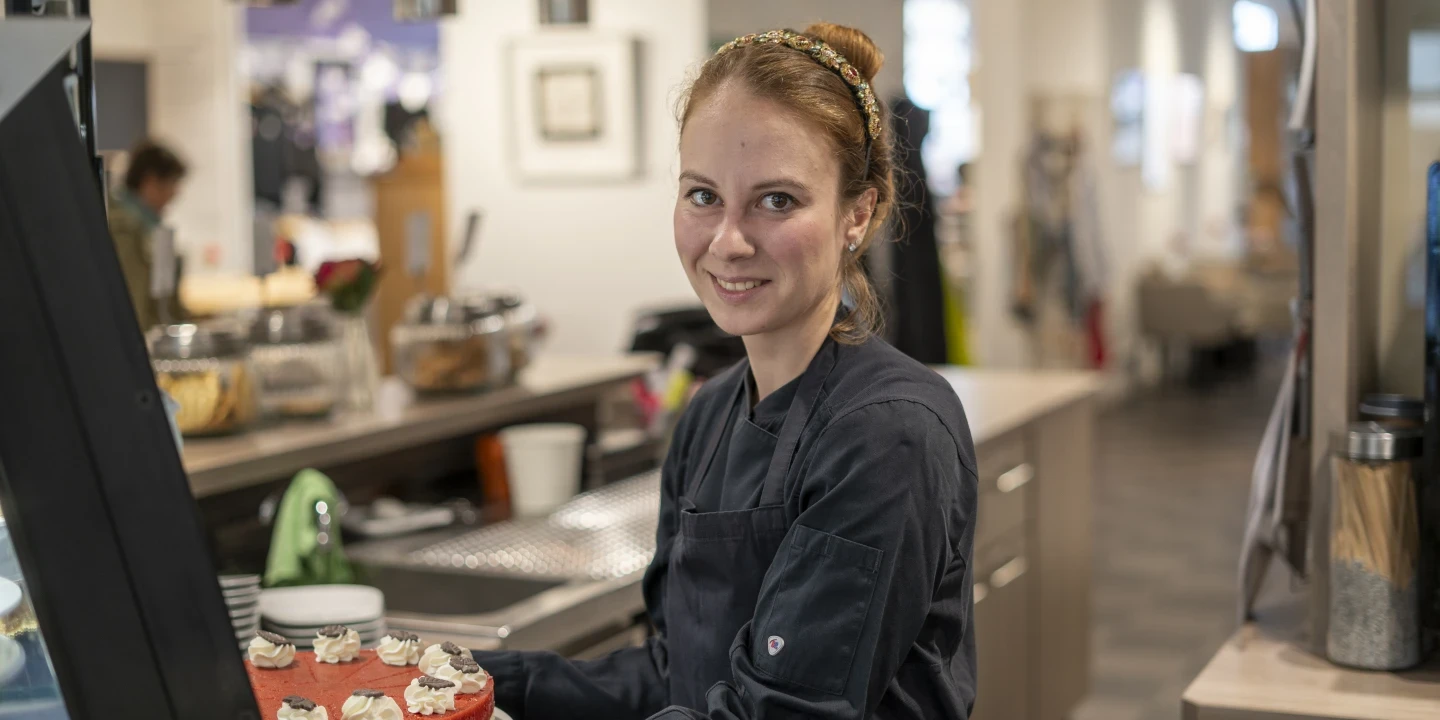 Frau in dunkelblauer Arbeitskleidung h&auml;lt einen Kuchen mit rotem Belag und Sahnetuffs in den H&auml;nden. Im Hintergrund Thekenbereich mit Gl&auml;sern, Bechern und weiteren Gegenst&auml;nden, sowie ein heller, stilvoll eingerichteter Raum.