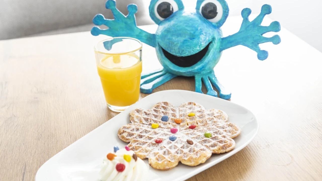 Blaues, humorvoll gestaltetes Froschmodell mit gro&szlig;en Augen und breitem L&auml;cheln im Hintergrund auf einem Holztisch. Im Vordergrund rechteckiger wei&szlig;er Teller mit herzf&ouml;rmiger Waffel, verziert mit bunten Smarties und Puderzucker, daneben ein Klecks Sahne mit weiteren Smarties. Links davon ein Glas Orangensaft.