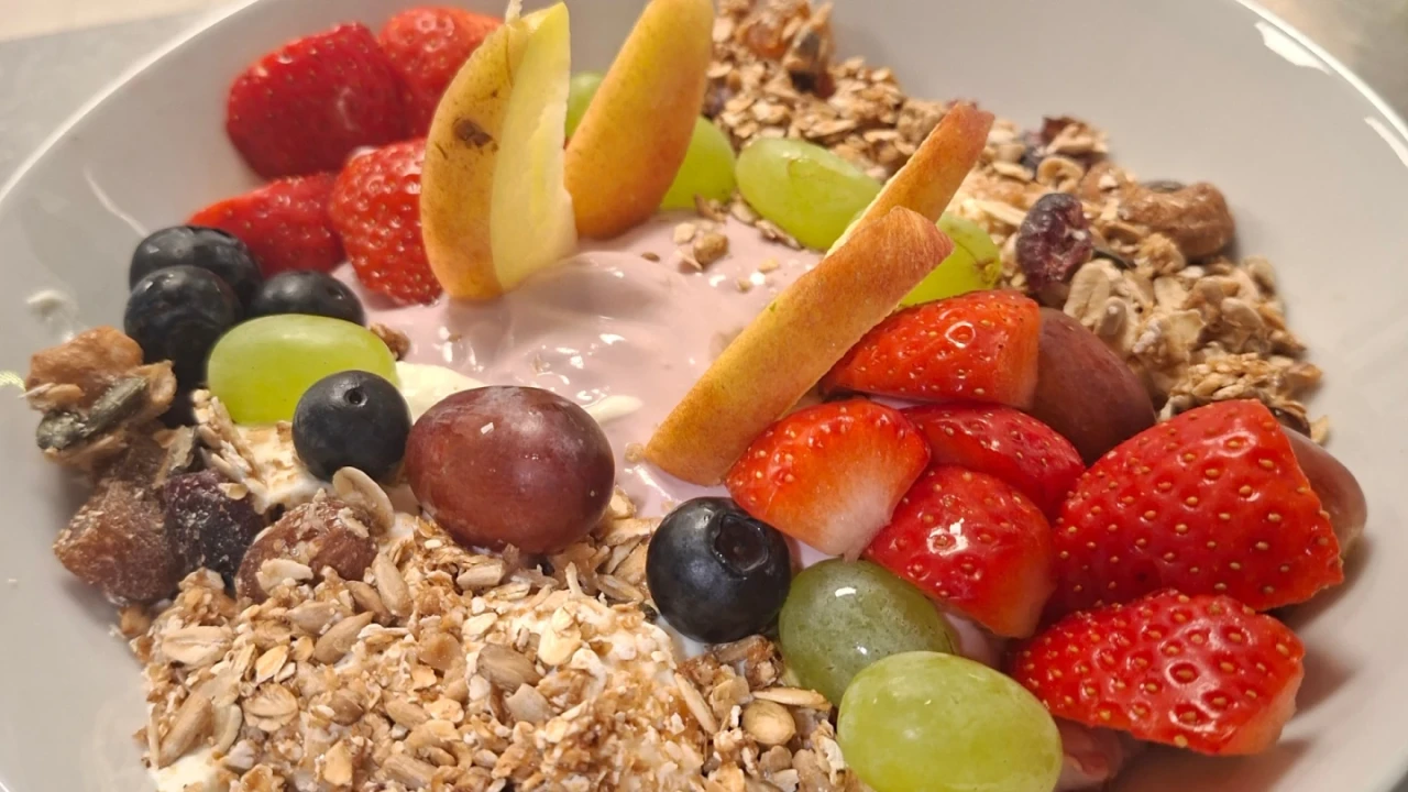 M&uuml;sli in einer wei&szlig;en Sch&uuml;ssel mit Joghurt in der Mitte, umgeben von Haferflocken, K&ouml;rnern, Rosinen und frischem Obst: geschnittene Erdbeeren, gr&uuml;ne und rote Weintrauben, Blaubeeren und Apfelscheiben vertikal arrangiert.
